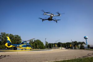 Medical helicopter that says Survival Flight in dark gold letters against dark blue background is on the ground at the Mcity Test Facility while a gray drone hovers overhead.